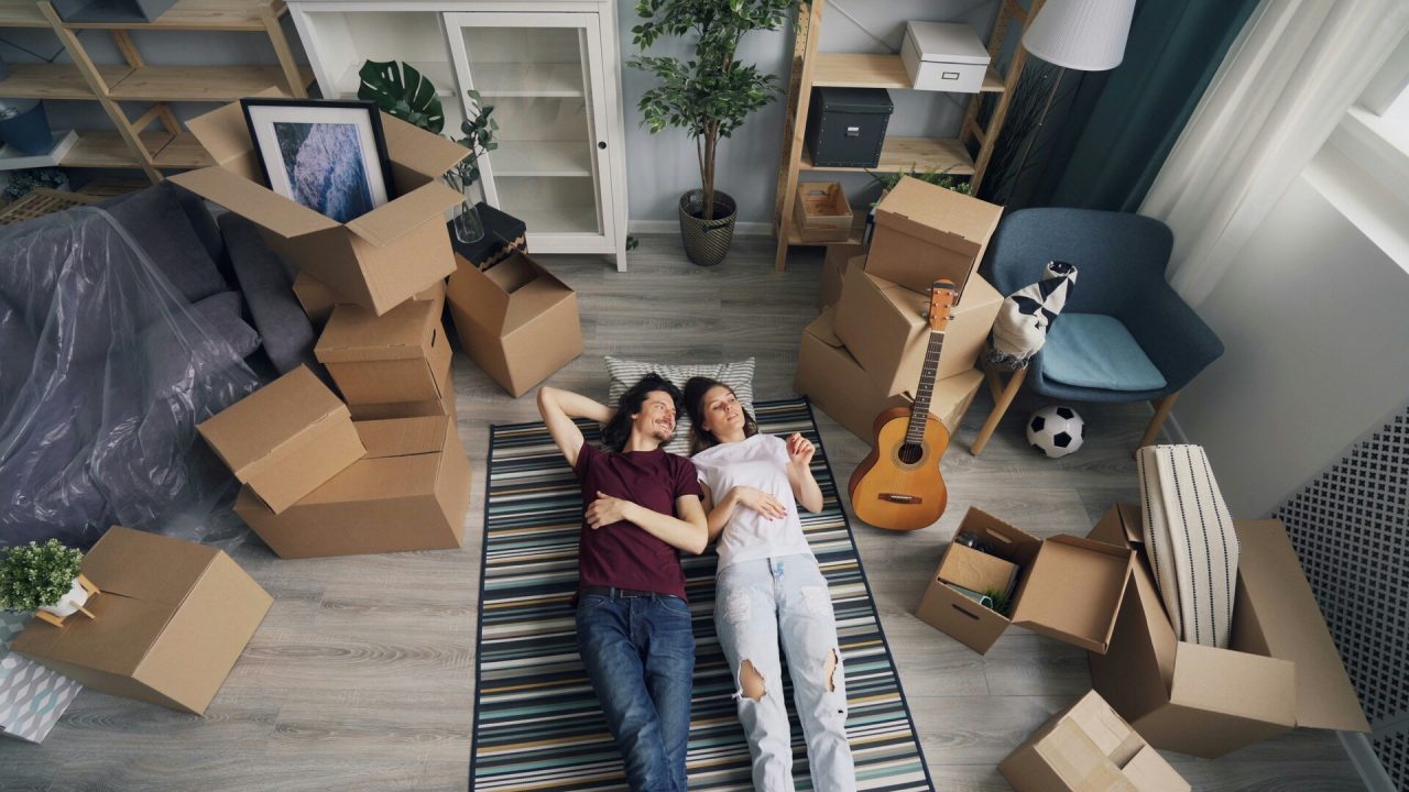 A happy couple lying on a rug in their new home, surrounded by cardboard moving boxes, looking relaxed and content.